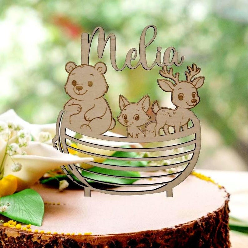 Pu&ograve; includere: Un cake topper in legno con il nome "Melia" in corsivo. Presenta un orso, una volpe e un cervo all'interno di un cestino di legno. Il design &egrave; pensato per essere utilizzato su una torta, con una finitura in legno naturale.