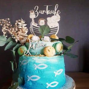 Pu&ograve; includere: Una torta a due piani con glassa blu, decorata con simboli di pesce bianchi, macarons e vegetazione. Un topper in legno recita "Zur Taufe" e presenta una giraffa, un leone e un elefante. Viene visualizzato anche il nome "Luca".
