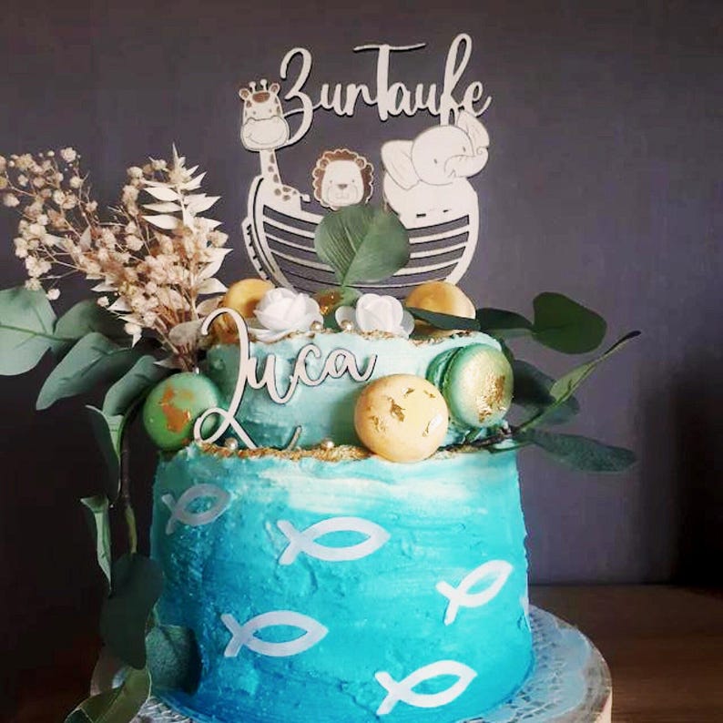 Pu&ograve; includere: Una torta a due piani con glassa blu, decorata con simboli di pesce bianchi, macarons e vegetazione. Un topper in legno recita "Zur Taufe" e presenta una giraffa, un leone e un elefante. Viene visualizzato anche il nome "Luca".
