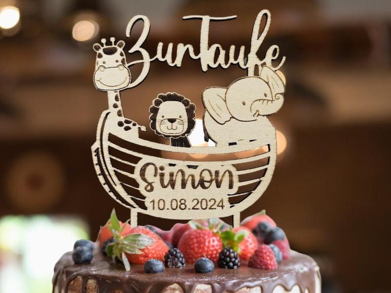 Pu&ograve; includere: Topper per torta in legno con le parole "Zur Taufe" e un design con giraffa, leone ed elefante. Sono inclusi anche il nome "Simon" e la data "10.08.2024". La torta &egrave; guarnita con fragole, mirtilli e more.