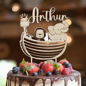 Pu&ograve; includere: Un cake topper in legno con il nome "Arthur" in corsivo. Il topper presenta una giraffa, un leone e un elefante in un design a barca. La torta &egrave; decorata con frutti di bosco freschi e glassa al cioccolato.