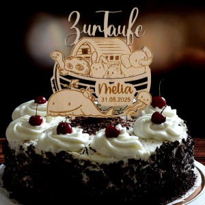 Pu&ograve; includere: Una torta decorata con glassa bianca, scaglie di cioccolato e ciliegie. Un cake topper in legno raffigura un'arca con animali, il nome "Melia" e la data "31.05.2025", con il testo tedesco "Zur Taufe" sopra.
