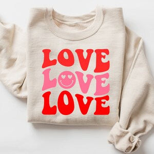 May include: A cream-colored sweatshirt with the word "LOVE" in red and pink, stacked vertically. The middle "LOVE" features a smiley face with heart eyes. The sweatshirt is folded neatly on a white surface.
