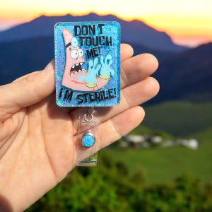 K&ouml;nnte beinhalten: Ein blauer und rosa Glitzer-Badgehalter mit einer Comicfigur, die blaue Handschuhe tr&auml;gt und den Text "Nicht anfassen! Ich bin steril!"