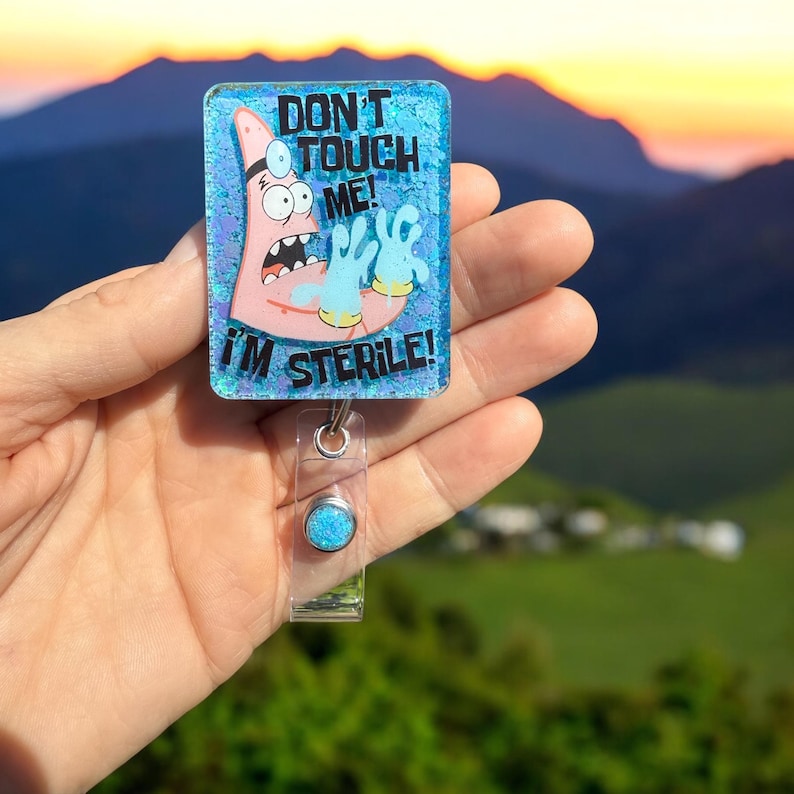 K&ouml;nnte beinhalten: Ein blauer und rosa Glitzer-Badgehalter mit einer Comicfigur, die blaue Handschuhe tr&auml;gt und den Text "Nicht anfassen! Ich bin steril!"