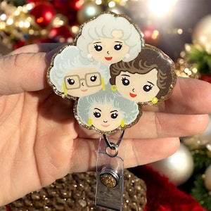 May include: A gold glitter badge reel with a clear plastic retractable cord and a round gold glitter charm featuring a cartoon image of three women with gray hair.