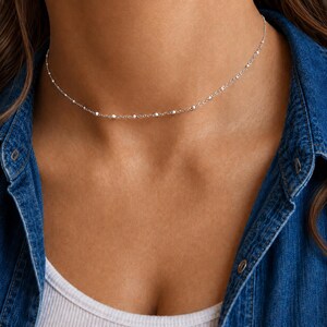 Könnte beinhalten: Eine zarte silberne Kettenhalskette mit kleinen, gleichmäßig verteilten Metallperlen entlang der gesamten Länge. Die Halskette liegt eng am Hals an, im Stil eines Chokers, und zeigt ein schlichtes, raffiniertes Design mit polierter Oberfläche.