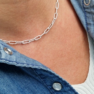 May include: A silver chain necklace with a delicate, textured link design. The necklace is worn by a person with a denim shirt and white top.