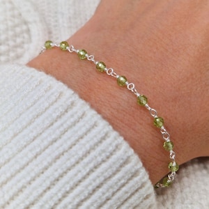 Könnte beinhalten: Ein zartes Armband aus Silberkette mit kleinen, facettierten grünen Edelsteinen. Das Armband wird am Handgelenk einer Person gezeigt.