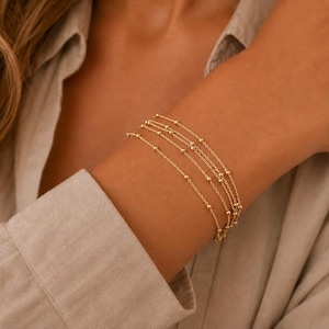Könnte beinhalten: Ein zartes goldenes Armband mit mehreren dünnen Ketten, die mit kleinen, gleichmäßig verteilten goldenen Perlen verziert sind. Das Design besteht aus mehreren geschichteten Strängen, die sich um das Handgelenk legen und ein raffiniertes Bild in poliertem Gold ergeben.