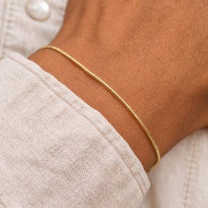 Pode incluir: Uma pulseira de corrente fina tipo cobra na cor dourada com uma textura metálica lisa, flexível e arredondada. A peça de joalharia apresenta um design de elos contínuos e uniformes que envolve o pulso, exibindo um acabamento polido.