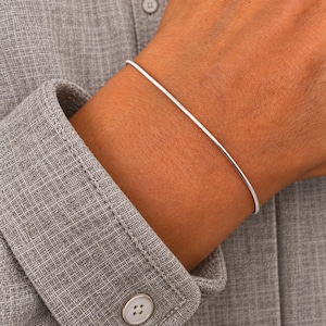 Könnte beinhalten: Ein dünner, glatter Armreif aus silberfarbenem Metall mit abgerundetem Profil. Das Armband ist ein einfacher, durchgehender kreisförmiger Ring, der auf der Haut aufliegt und eine polierte, reflektierende Oberfläche aufweist.