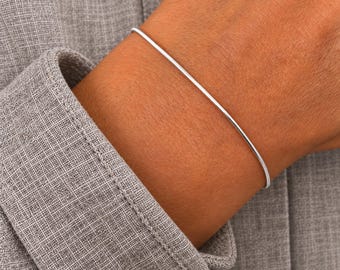 May include: A thin, smooth, silver-toned metal bangle bracelet with a rounded profile. The bracelet is a simple, continuous circular band that rests against the skin, showcasing a polished, reflective surface and a clean, minimalist design.