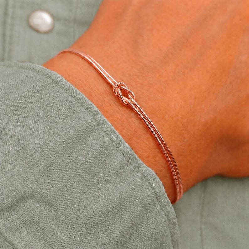 Puede incluir: Una pulsera de oro rosa con un dise&ntilde;o de cadena fina y delicada con un detalle de nudo central. La pulsera est&aacute; fabricada con un material met&aacute;lico fino y flexible que se envuelve alrededor de la mu&ntilde;eca, mostrando un acabado pulido y una est&eacute;tica sencilla.