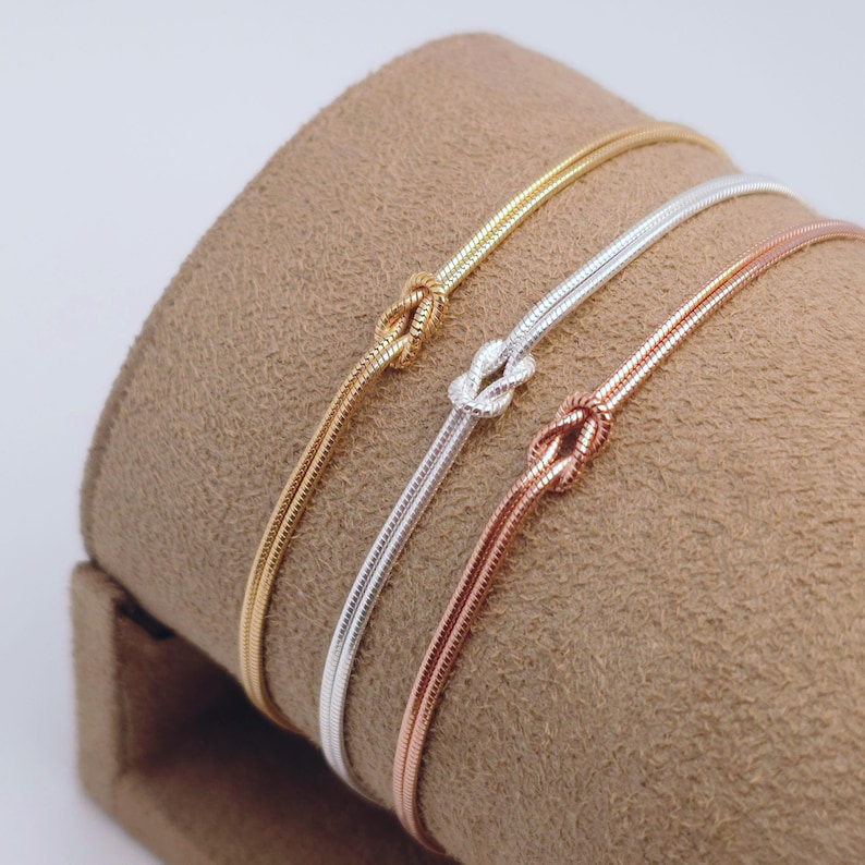 Puede incluir: Tres pulseras finas de cadena de serpiente en tonos oro, plata y oro rosa. Cada pulsera presenta un detalle de nudo central. Las cadenas tienen una textura suave y flexible, dispuestas una al lado de la otra para mostrar sus acabados.