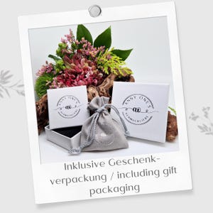 May include: Two white square jewelry boxes and a small grey fabric drawstring pouch, each featuring the black circular logo Anny One's Schmuckliebe. The items are arranged with a bouquet of pink and green flowers and a piece of driftwood.
