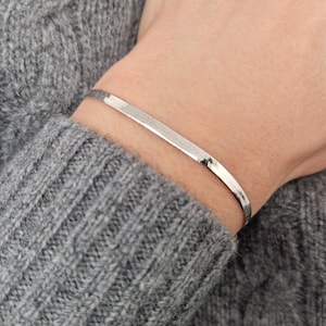 Op de afbeelding: Een zilveren kettingarmband met een plat, rechthoekig ontwerp. De armband wordt getoond op de pols van een persoon.
