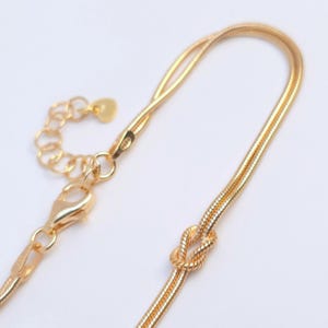 Puede incluir: Un collar de cadena de serpiente en tono dorado con un peque&ntilde;o detalle de nudo decorativo. El collar incluye un cierre de mosquet&oacute;n, una cadena extensora corta con eslabones circulares y un peque&ntilde;o dije plano en forma de coraz&oacute;n al final.