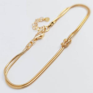 Puede incluir: Una pulsera de cadena de serpiente en tono dorado con un detalle de nudo central. La pulsera incluye un cierre de mosquet&oacute;n y una cadena extensora ajustable con un peque&ntilde;o dije circular y plano en el extremo, logrando un aspecto pulido.