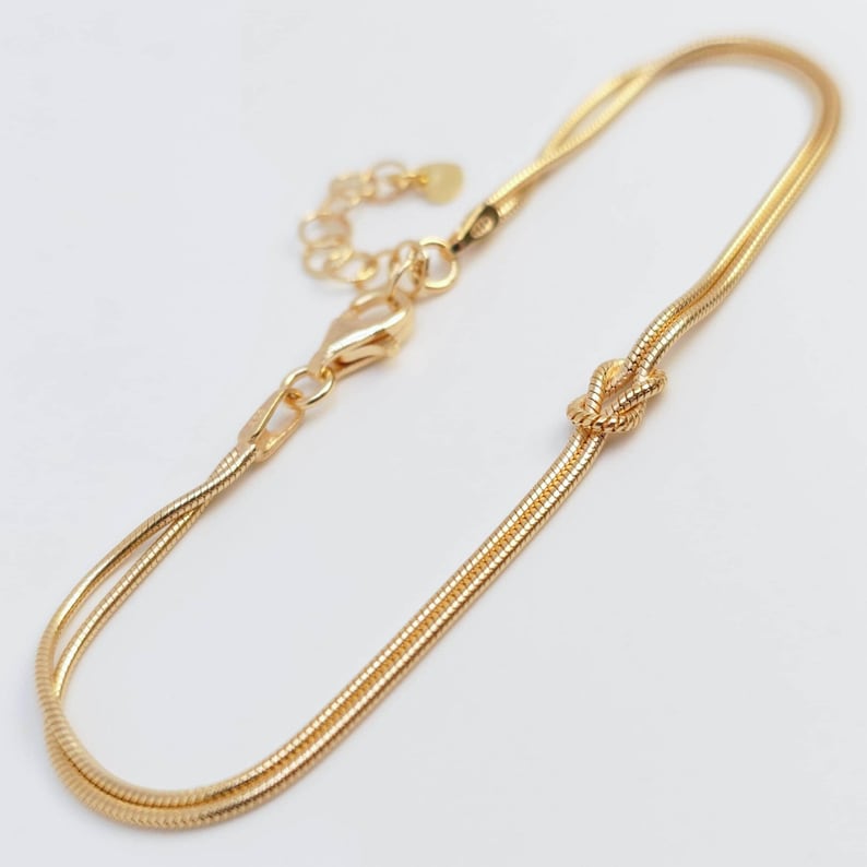 Puede incluir: Una pulsera de cadena de serpiente en tono dorado con un detalle de nudo central. La pulsera incluye un cierre de mosquet&oacute;n y una cadena extensora ajustable con un peque&ntilde;o dije circular y plano en el extremo, logrando un aspecto pulido.
