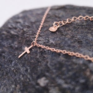 May include: A rose gold-toned necklace featuring a delicate chain and a small, simple cross pendant. The chain includes a short extender section with a small, flat, heart-shaped charm at the end, showcasing a polished metallic finish throughout.