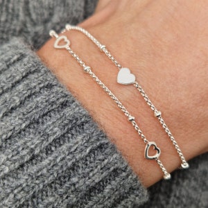 Könnte beinhalten: Zwei silberne Armbänder mit Herz-Anhängern und kleinen silbernen Perlen. Die Armbänder sind übereinander am Handgelenk einer Person gelegt.
