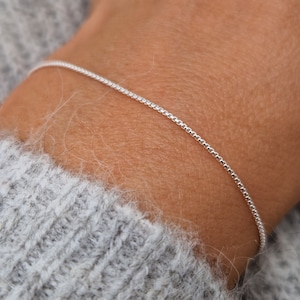 Peut inclure: Un bracelet en argent fin avec une chaîne de type boîte. Le bracelet est présenté sur un poignet, avec un pull gris en arrière-plan. La chaîne est fine et composée de petits maillons carrés.