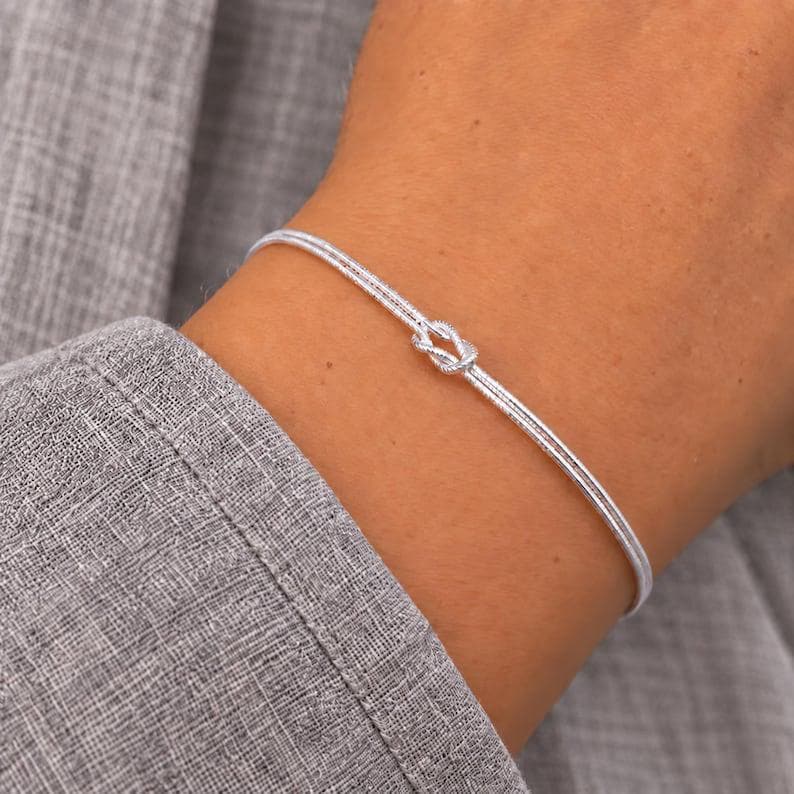 Puede incluir: Una pulsera r&iacute;gida fina de tono plateado pulido con un dise&ntilde;o de nudo central. La pulsera tiene una estructura de alambre liso y redondeado con un acabado met&aacute;lico que refleja la luz, creando una apariencia sencilla y limpia.
