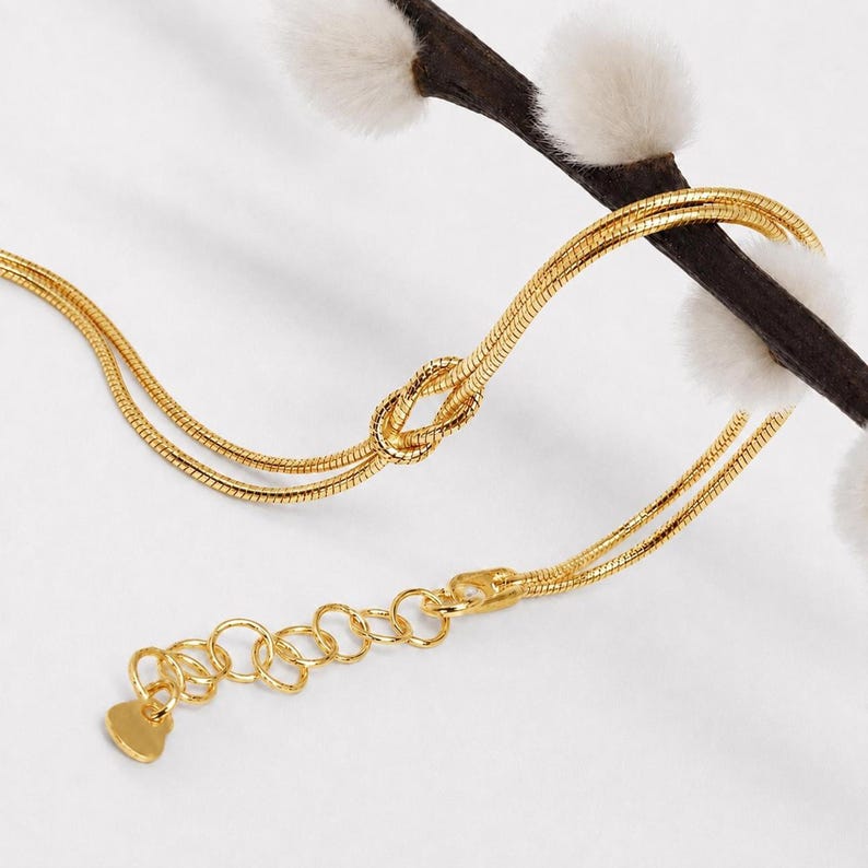 Puede incluir: Una pulsera de cadena de serpiente dorada con un nudo central. La pulsera incluye una cadena extensora ajustable con eslabones circulares y un peque&ntilde;o dije plano en forma de coraz&oacute;n en el extremo, colocada sobre una rama oscura con brotes blancos.