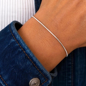 Peut inclure: Un bracelet fin et flexible en chaîne serpent de couleur argentée porté autour du poignet. Le bracelet présente une surface métallique lisse et continue avec une finition polie, créant un aspect élégant et discret sur la peau.