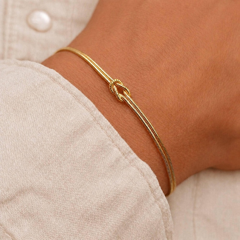Puede incluir: Una pulsera fina de oro con un dise&ntilde;o delicado de cadena de serpiente y un nudo central. El acabado dorado met&aacute;lico refleja la luz, resaltando la textura suave y flexible de la cadena mientras rodea la zona de la mu&ntilde;eca.