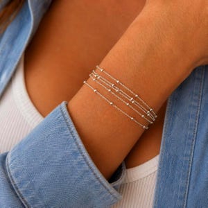 May include: A delicate silver bracelet featuring multiple thin chains adorned with small, evenly spaced metallic beads. The design consists of several layered strands that wrap around the wrist, creating a refined and uniform appearance in polished silver.
