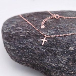 May include: A rose gold-toned necklace featuring a delicate chain and a small, simple cross pendant. The chain includes an adjustable section with a small circular tag at the end, showcasing a refined and minimalist design for everyday wear.