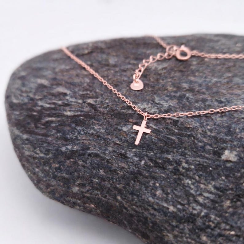 May include: A rose gold-toned necklace featuring a delicate chain and a small, simple cross pendant. The chain includes an adjustable section with a small circular tag at the end, showcasing a refined and minimalist design for everyday wear.