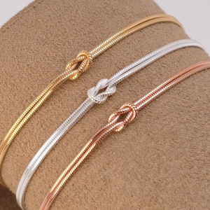 Puede incluir: Tres pulseras finas de cadena de serpiente en tonos oro, plata y oro rosa. Cada pulsera presenta un detalle de nudo central formado por la propia cadena. Las cadenas met&aacute;licas tienen una textura suave y flexible con acabado pulido.