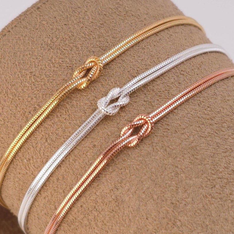 Puede incluir: Tres pulseras finas de cadena de serpiente en tonos oro, plata y oro rosa. Cada pulsera presenta un detalle de nudo central formado por la propia cadena. Las cadenas met&aacute;licas tienen una textura suave y flexible con acabado pulido.