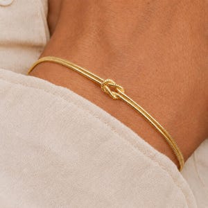Puede incluir: Una pulsera de tono dorado que presenta un dise&ntilde;o de cadena fina y delicada con un detalle de nudo central. La pulsera est&aacute; construida con dos hilos paralelos que se unen en el nudo, creando un bucle continuo y refinado alrededor de la mu&ntilde;eca.