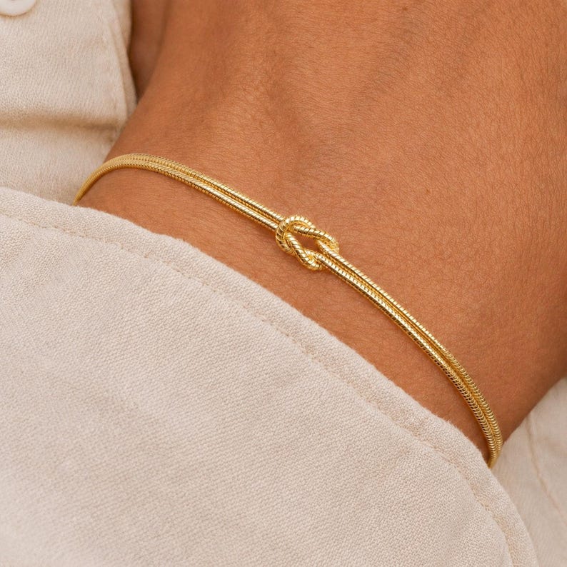 Puede incluir: Una pulsera de tono dorado que presenta un dise&ntilde;o de cadena fina y delicada con un detalle de nudo central. La pulsera est&aacute; construida con dos hilos paralelos que se unen en el nudo, creando un bucle continuo y refinado alrededor de la mu&ntilde;eca.