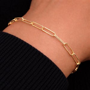 May include: A gold chain bracelet featuring elongated, rectangular links connected in a repeating pattern. The bracelet has a polished, metallic finish and is designed with a simple, continuous structure that wraps around the wrist area.