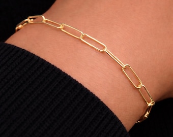 Puede incluir: Una pulsera de cadena dorada con eslabones rectangulares alargados conectados en un patrón repetitivo. La pulsera tiene un acabado metálico pulido y está diseñada con una estructura simple y continua que rodea la zona de la muñeca.