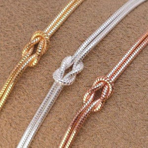 Puede incluir: Tres cadenas de serpiente en tonos oro, plata y oro rosa, cada una con un nudo decorativo atado en el centro. Las cadenas tienen una textura suave, flexible y redondeada, mostrando un acabado met&aacute;lico en los tres colores distintos.