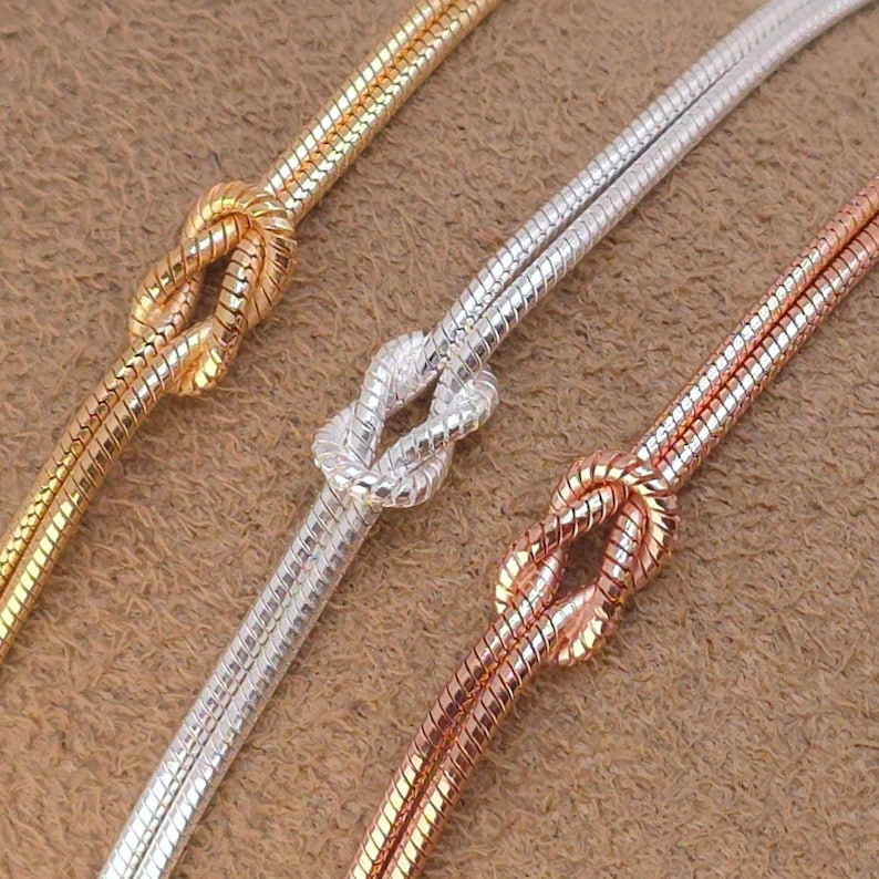 Puede incluir: Tres cadenas de serpiente en tonos oro, plata y oro rosa, cada una con un nudo decorativo atado en el centro. Las cadenas tienen una textura suave, flexible y redondeada, mostrando un acabado met&aacute;lico en los tres colores distintos.