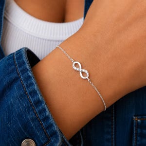 Peut inclure: Un bracelet à chaîne de couleur argentée doté d'un pendentif central en forme de symbole de l'infini. Le symbole est orné de petites pierres claires et scintillantes incrustées le long de ses courbes. La chaîne est fine et polie.