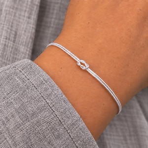 Könnte beinhalten: Ein dünner, silberfarbener Armreif mit einem zentralen Knotendetail. Das Armband hat eine texturierte, schimmernde Oberfläche und eine zarte, schlichte Konstruktion, die sich um das Handgelenk legt und eine durchgehende, raffinierte kreisförmige Schlaufe bildet.