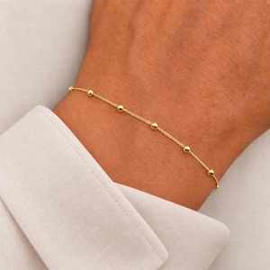 Könnte beinhalten: Ein zartes Goldkettenarmband mit gleichmäßig verteilten kleinen Goldperlen entlang der Kette. Das Armband wird um das Handgelenk getragen und zeigt ein schlichtes, raffiniertes Design mit einer polierten metallischen Oberfläche und feinen Gliedern.