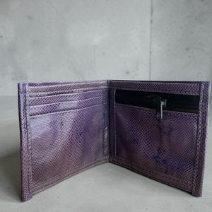 May include: Open purple bi-fold wallet with a textured, snake-like pattern. The wallet has multiple card slots and a zipped pocket. The interior is black. The wallet is open, displaying its interior.