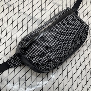 May include: A black and white checkered fanny pack with a black zipper and strap. The bag is rectangular and has a grid pattern. The strap is adjustable and made of black material.