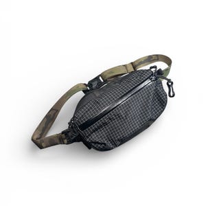 Peut inclure: Sac banane noir et blanc à motif quadrillé avec une sangle camouflage. Le sac a un compartiment principal zippé et une tirette noire. La sangle est réglable et comporte une boucle en plastique noir.