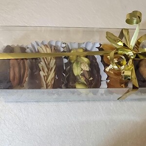 May include: A clear plastic box filled with six individually wrapped chocolate-covered treats. The treats are arranged in a row and include a variety of fillings, such as nuts, dried fruit, and pistachios. The box is tied with a gold ribbon and bow.