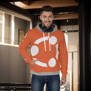 Puede incluir: Sudadera con capucha naranja con un diseño gráfico blanco y forro de capucha gris. La sudadera tiene un bolsillo de canguro y cordones blancos. Las mangas tienen un pequeño diseño blanco. La persona lleva vaqueros azules y auriculares.